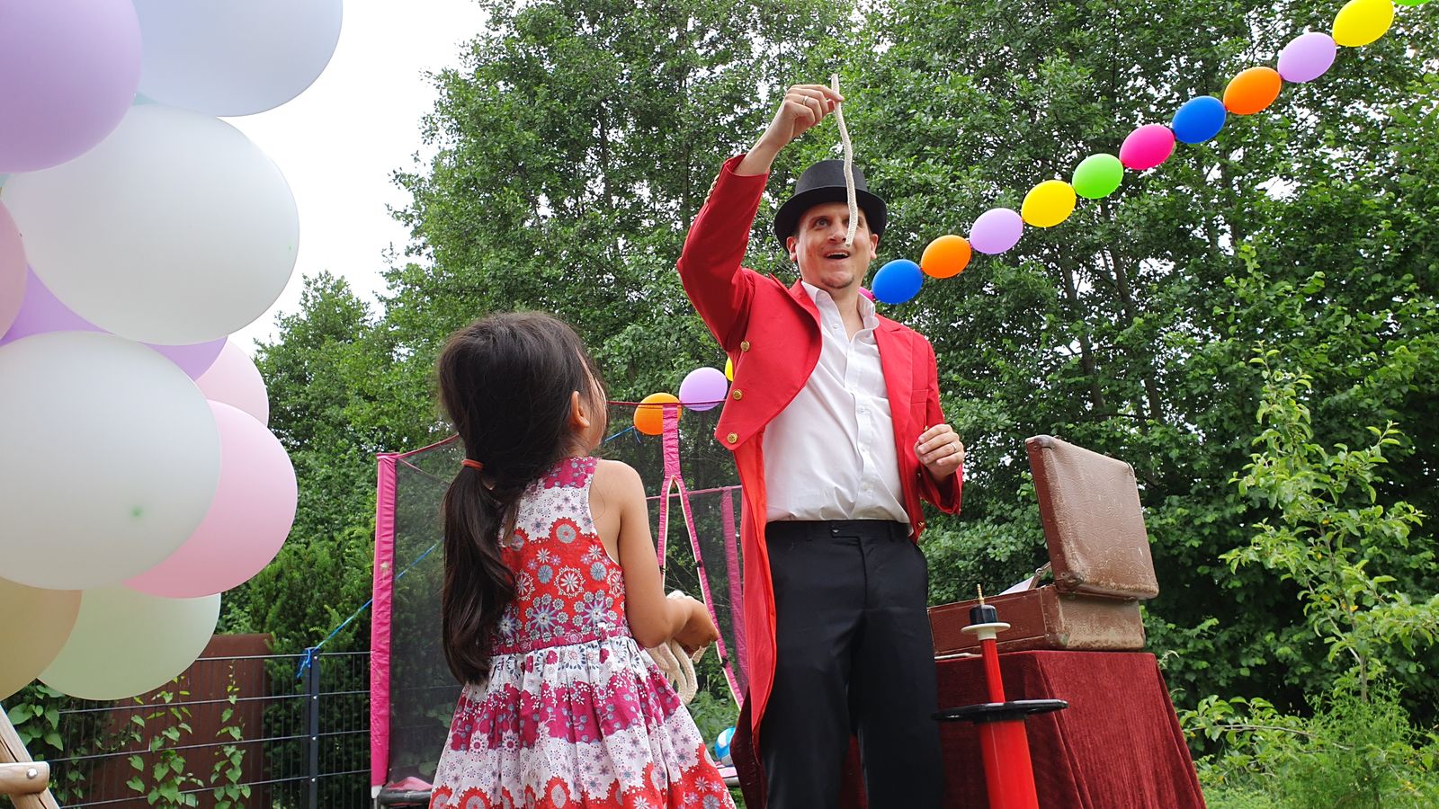 Mr. Hagen im roten Frack zaubert im Garten mit einem Seil, ein Kind schaut zu, überall Luftballons
